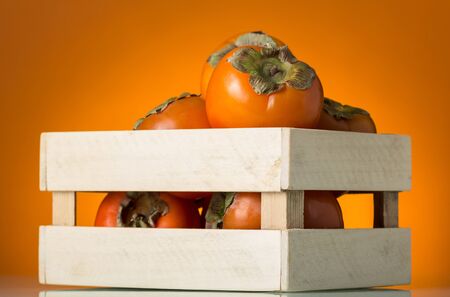 Wooden box full of ripe persimmon fruit on bright orange backgroundの写真素材