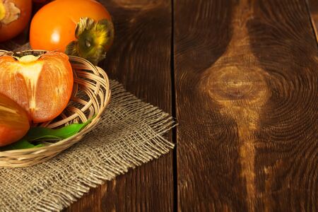 Persimmon and piece of fruit in wicker basket on burlap, on dark wooden surfaceの写真素材