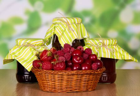 Basket of ripe strawberries, raspberries and cherries, jars of jam on abstract green background.の写真素材
