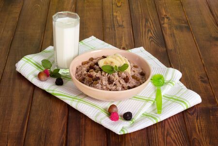 A bowl of porridge, a glass of milk, a spoon, nuts, raspberries and a kitchen towel on dark wooden background.の写真素材