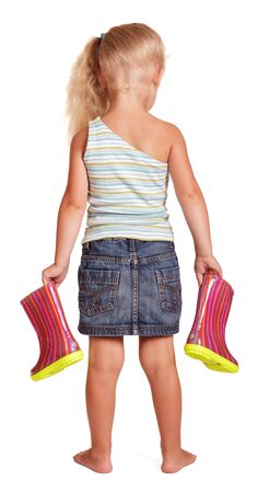 Little blond girl in a skirt and blouse stand holding rubber boots isolated on white background. Back view.の写真素材