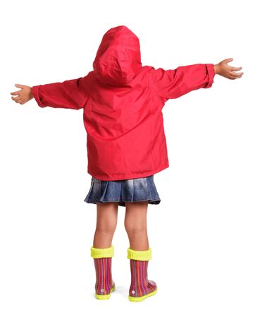 Little girl in a red jacket, blue skirt and rubber boots standing with arms outstretched isolated on white background. Back view.の写真素材