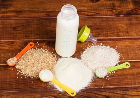 Bottle of infant formula, a heap of different cereals and measuring spoon against a background of dark wood.の写真素材