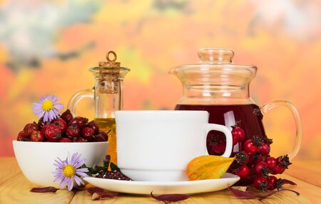 A bowl of dog rose berries, drink a jug, a cup of tea and a bottle of oil on a background of autumn leaves.の写真素材