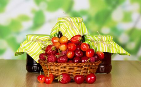 Ripe strawberries, raspberries and cherries in a basket, jars of jam on abstract green background.の写真素材