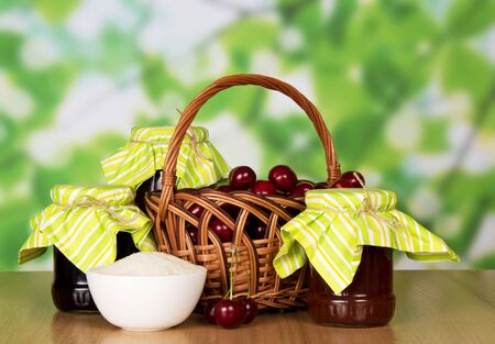 Jars of jam, a basket of cherries and a cup of sugar on abstract green background.の写真素材