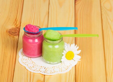 Glass jars with vegetables, fruit puree, plastic spoon and daisy on the background of light wood.の写真素材