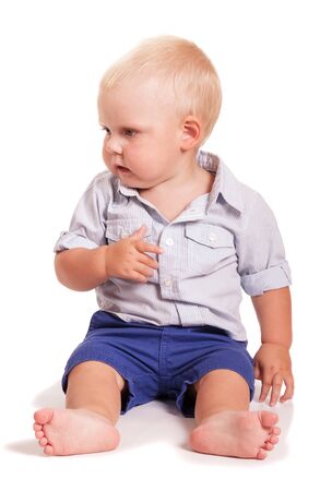 A little boy in a bright shirt with stripes and blue shorts sitting isolated on white background.の写真素材