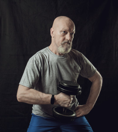 An elderly shaven-headed man with a gray beard trains his arms with dumbbellsの写真素材