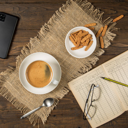 Morning coffee - a cup, a notebook, a pencil, glasses and a smartphone on a brown wooden table with a napkinの写真素材