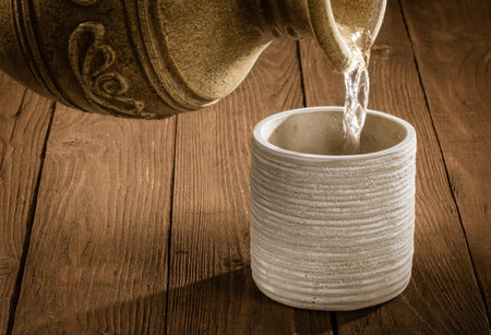 A jet of water pours from an earthenware jug into an earthenware glass standing on a wooden tableの写真素材