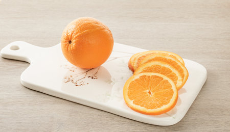 A whole orange and orange slices lie on a white ceramic kitchen board
