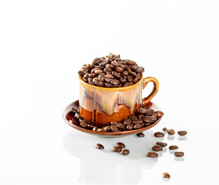 A ceramic cup filled to the brim with coffee beans stands on a white mirror background. Coffee beans are scattered around the cup. The cup is beautifully reflected in the mirror backgroundの写真素材