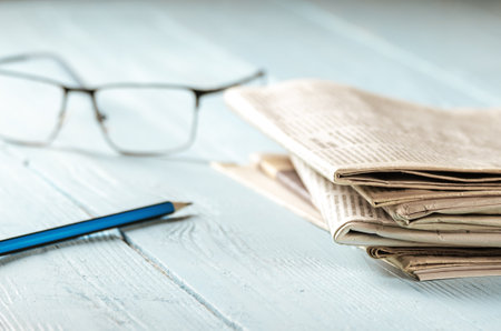 Reading the morning news. A stack of newspapers, glasses and a pencil on a wooden table lit by the morning sunの写真素材