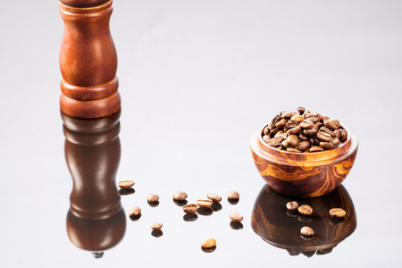 A wooden cup filled with coffee beans is reflected in a black mirror background. A beautiful contrasting reflection, scattered coffee beans and a wooden coffee grinder in the backgroundの写真素材
