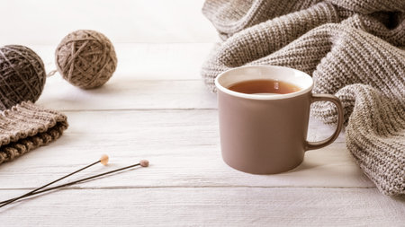 A close-up of a cozy cup of tea surrounded by knitted items and wool yarn balls, creating a warm and...