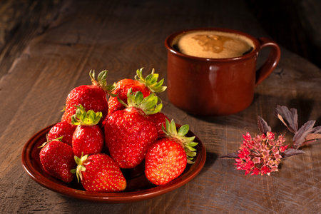 This image features a vibrant saucer filled with ripe, juicy strawberries alongside a steaming cup of coffee topped with frothy foam, beautifully presented on a rustic wooden table.の写真素材