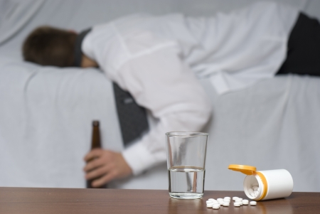 Glass of water and pills on the table. In the background out of focus a passed out businessman on the sofa.の写真素材