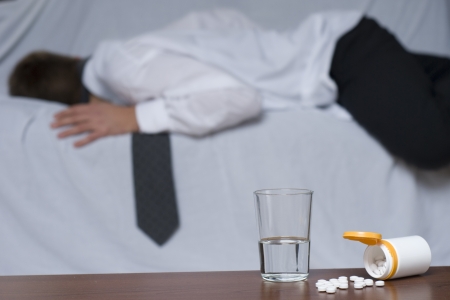 Glass of water and pills on the table. In the background out of focus a passed out businessman on the sofa.の写真素材
