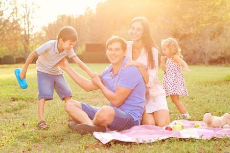 Happy family having a picnic in the park on nature. Father, mother and kids playing on the grass in the park in the evening sunlight. Parents and children laughing, smiling, playing and having fun together.の写真素材