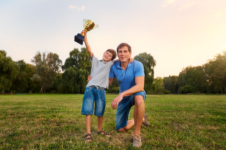 The son of a champion with a gold cup champion with his father in the park embracing outdoors in nature smiling, laughing, fun, joy.の写真素材
