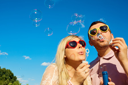 Concept of happiness, cheerful, fun, love. Happy couple in sunglasses hearts having fun blowing soap bubbles against the blue sky.の写真素材