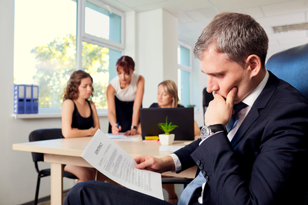 The concept of making business solution. Serious businessman reading a contract sitting in the chair in the office on the background of colleagues, partners and clients working at the table at laptop.の写真素材