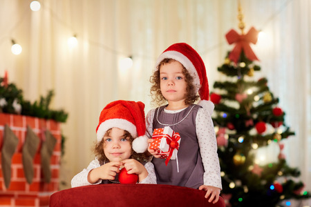 Sisters twins child in Christmas hats Santa with gifts near the Christmas tree indoors.の写真素材