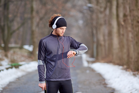 Man in sportswear jogging wearing headphones looking at his watch on the hand in park autumn spring winter.の写真素材