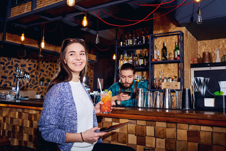 A girl with  cocktail smiles with tablet behind counter at the bar.の写真素材