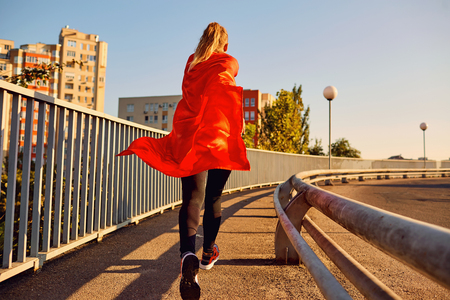 Woman super-hero runner in a red raincoat jogs on a bridge in the city.の写真素材