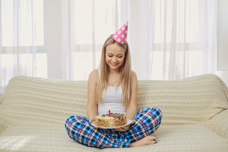A blonde girl in a pajama cap with a cake with a candle celebrating a birthday sitting on the couch in the room on the window background.の写真素材