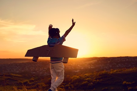 A happy boy in a superhero costume is playing with an airplane on the nature at sunset.の写真素材