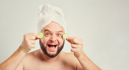 A funny fat man with a beard with a cucumber mask in the spa salon.の写真素材