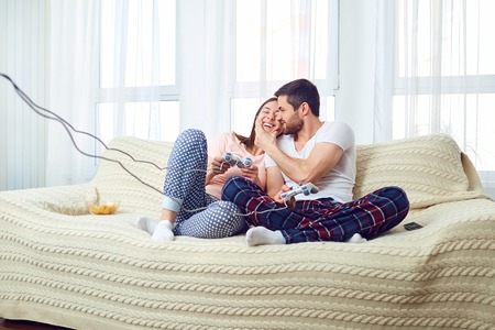 Couple playing video game sitting on sofa in room on window background.
