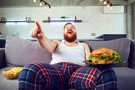 Thick funny man in pajamas with a burger sitting on the couch.の写真素材