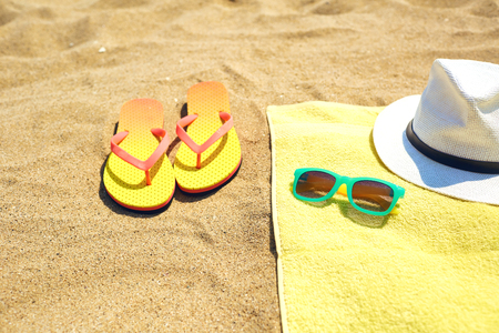 Trendy flip flops placed on sand near yellow towel with stylish shades and hat on sunny day on beachの写真素材