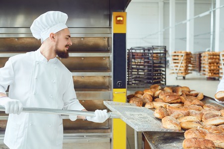 A baker with a shovel takes fresh bread from the oven in the bakery.の写真素材