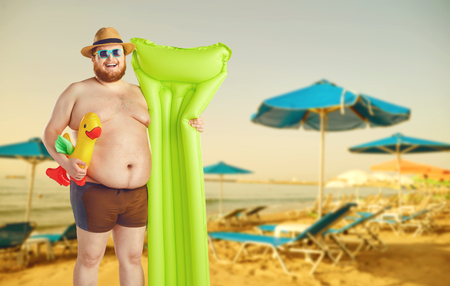Fat funny man with a beard in a hat in swimming trunks with an inflatable mattress on a gray background. Summer seaside concept. Vacation.の写真素材