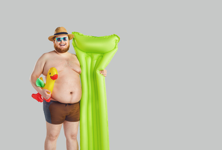 Fat funny man with a beard in a hat in swimming trunks with an inflatable mattress on a gray background. Summer seaside concept. Vacation.の写真素材