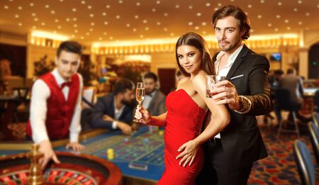 Beautiful glamour couple with glasses of alcohol in hand are standing against the background of casino poker roulette.の写真素材