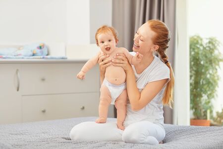Happy mother smiles at baby sitting on bed in room. Motherhood. Mothers Day.の写真素材