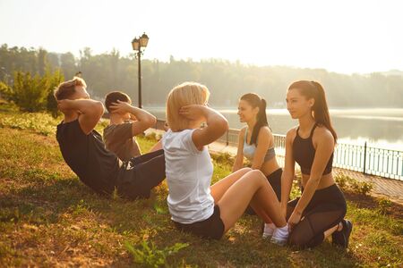 A group of people doing exercises sitting on the grass in the summer park in autumn. Athletes do abs...