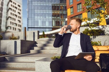 A bearded businessman is drinking coffee while sitting on a city street against the background of a business building.の写真素材