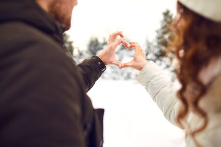 Crop back view of couple stacking hands in heart symbol while standing close in winter woods