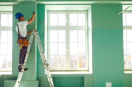A builder standing on a ladder installs drywall at a construction site inside an office.