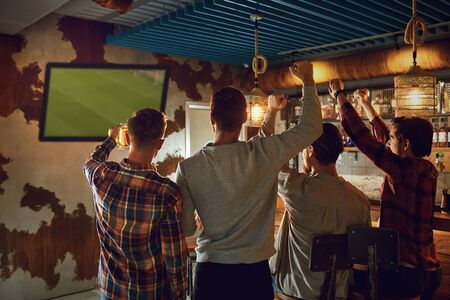 A group of friends watching tv football in a sports bar. Back view. Fans cheerfully cheer in a...