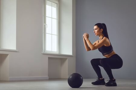 Squat exercises. Athletic brunette girl in black sportswear with dumbbells in her hands doing squats in a room indoors.の写真素材