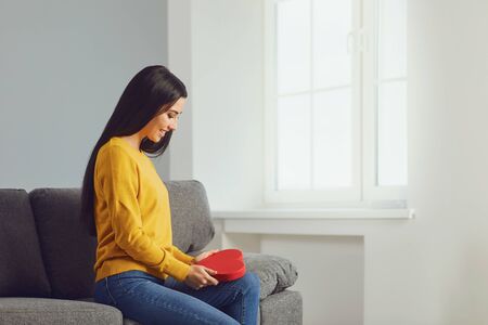 Beautiful girl holding a red heart in her hands while sitting on a sofa in a gray room. Valentines day.の写真素材
