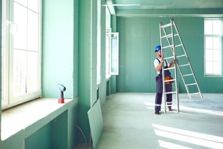 A builder standing on a ladder installs drywall at a construction site inside an office.の写真素材